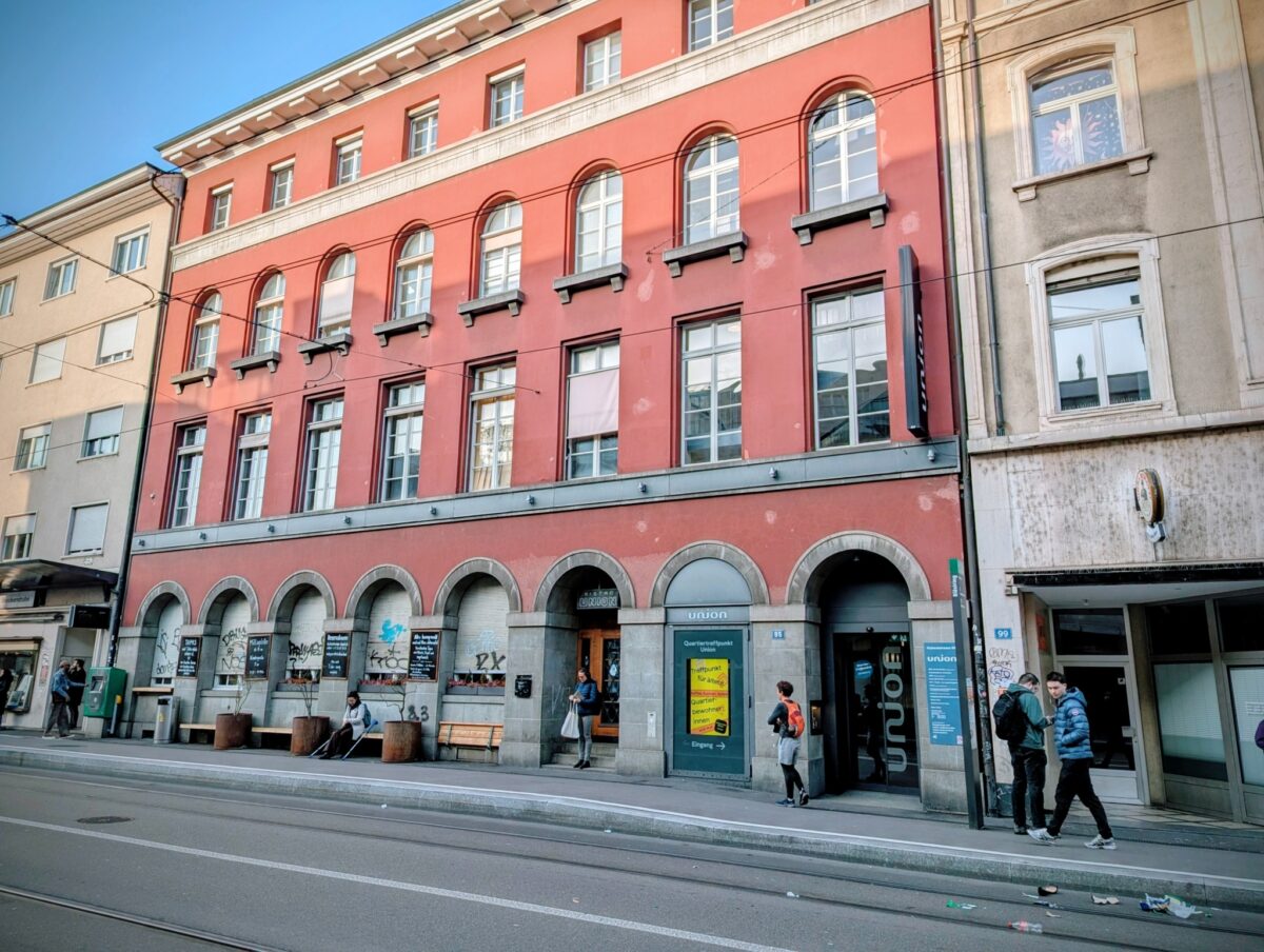 Ein rotes Gebäude mit mehreren Arkaden und Passanten auf der Straße.
