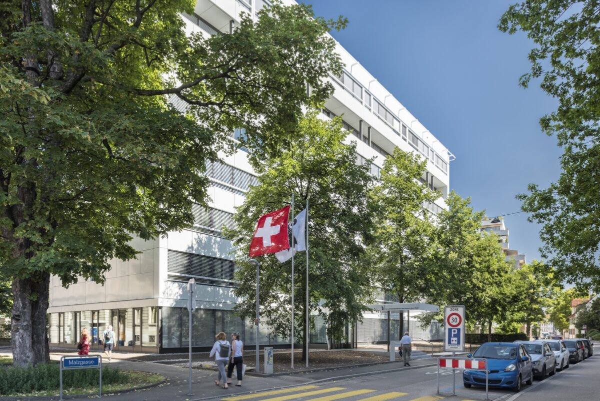 Ein modernes Gebäude mit Schweizer Flagge vor einem Baum und einer Straße.
