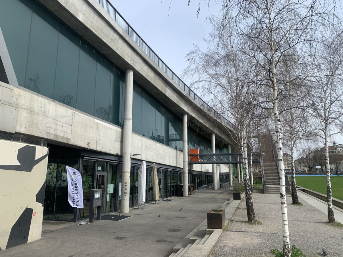 Moderne Gebäude mit großen Glasfenstern und Betonpfeilern, umgeben von kahlen Bäumen.