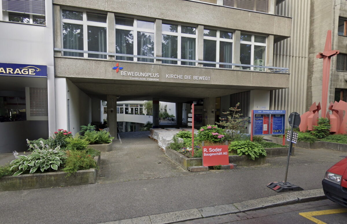 "Eingang eines Gebäudes mit Schildern und bepflanzten Blumenbeeten."