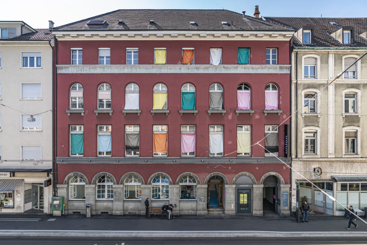Ein rotes Gebäude mit bunten Tüchern in den Fenstern und Menschen auf der Straße.