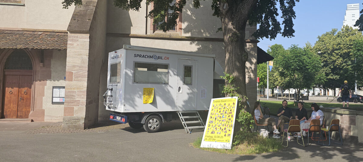 Ein weißer Sprachmobil-Wagen steht vor einem Gebäude mit Menschen auf Bänken daneben.