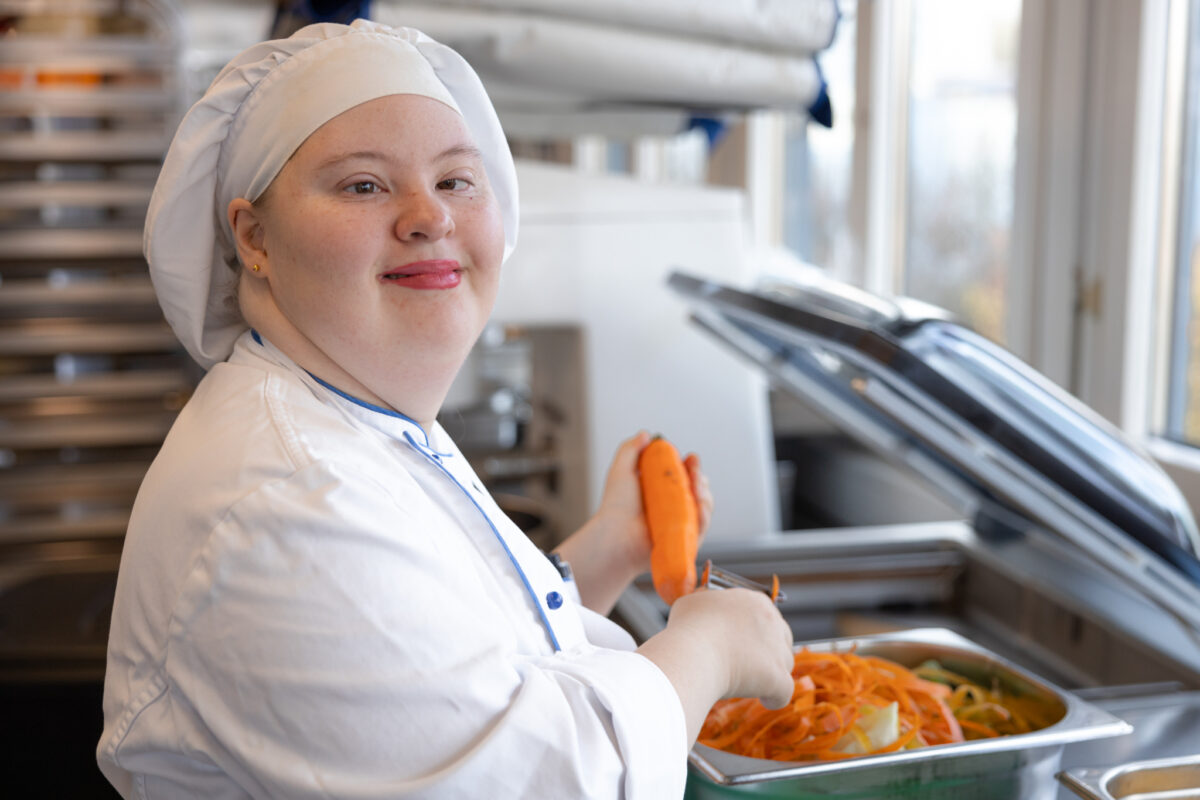 Eine Köchin in weißer Uniform serviert Gemüse in einer Küche.
