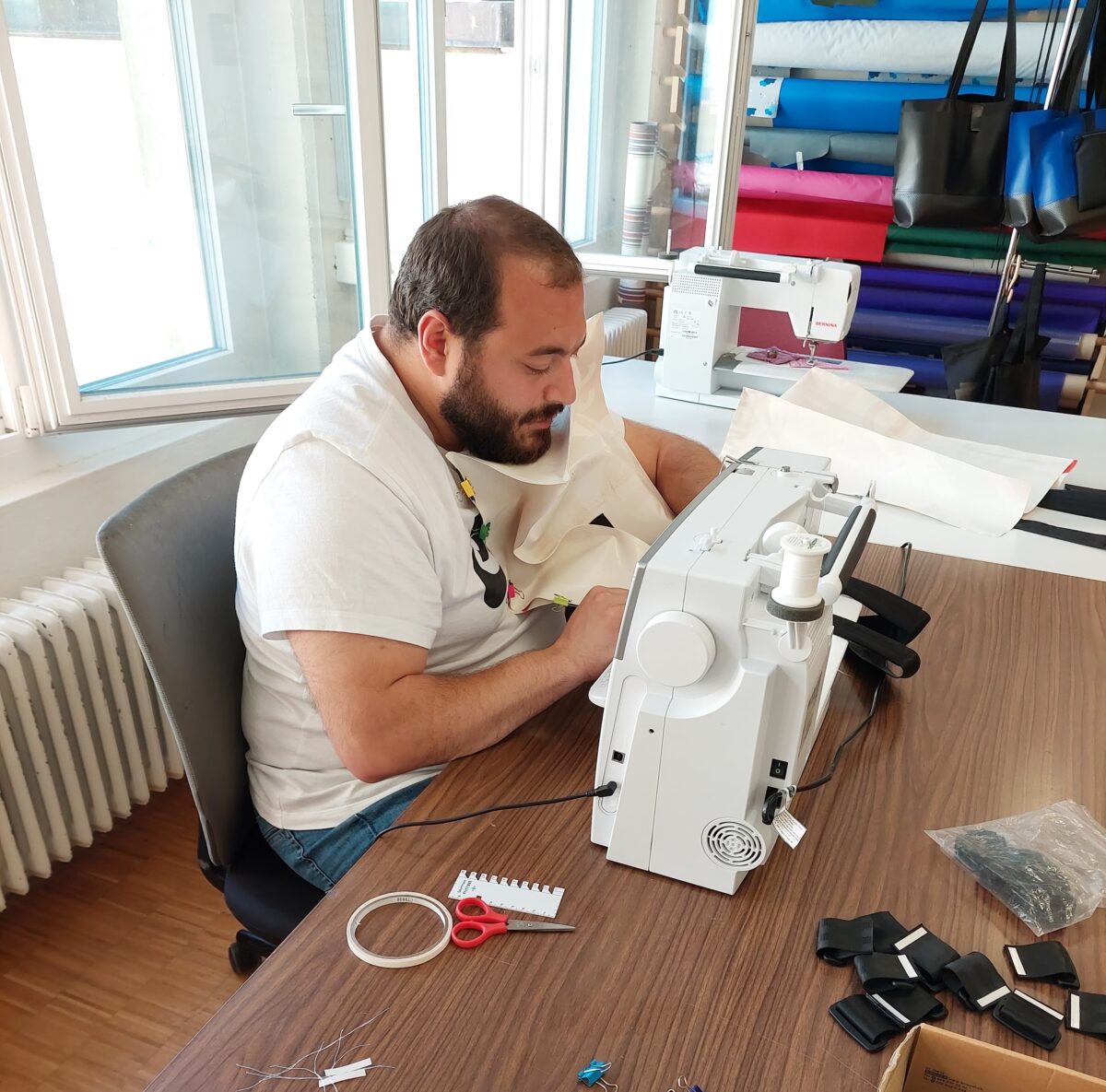 Ein Mann arbeitet konzentriert an einer Nähmaschine an einem Tisch mit Nähutensilien.