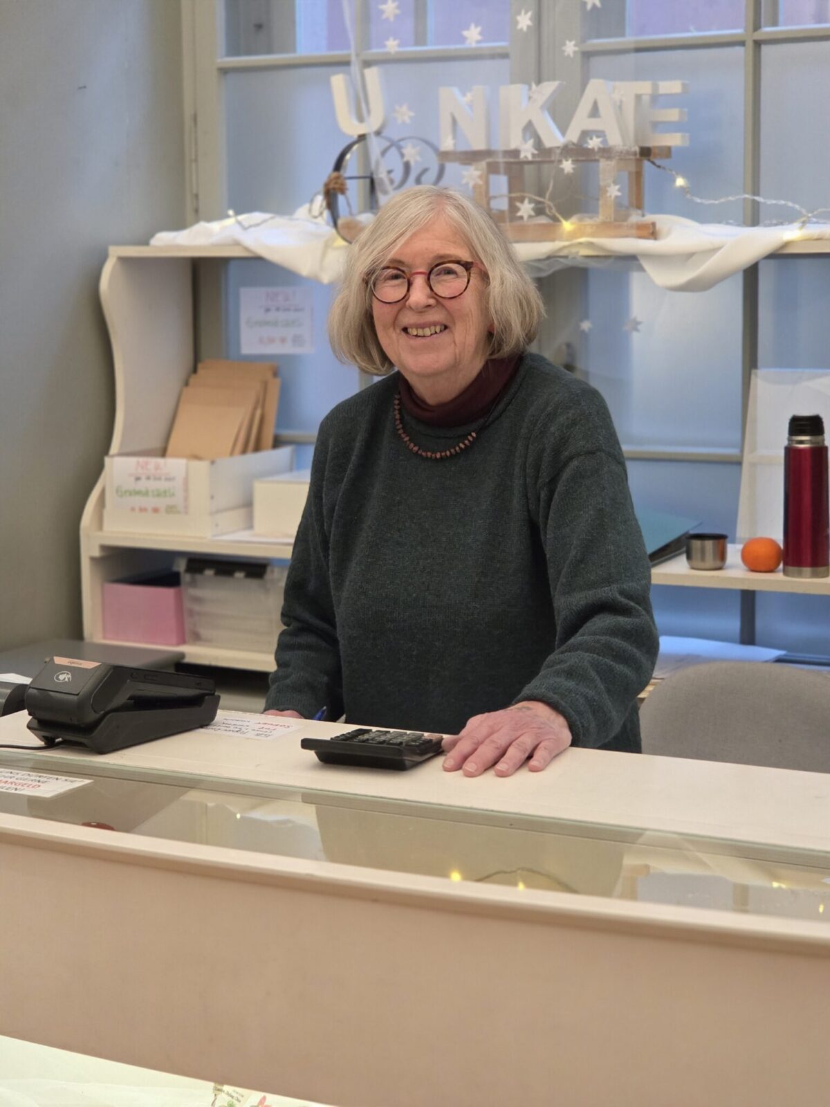 Eine lächelnde Frau steht hinter einem Verkaufstresen in einem Geschäft.