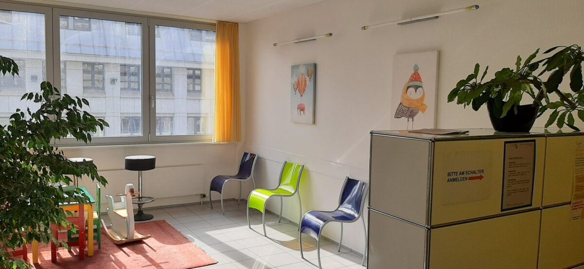 Ein heller Warteraum mit bunten Stühlen, Pflanzen und Bildern an der Wand.