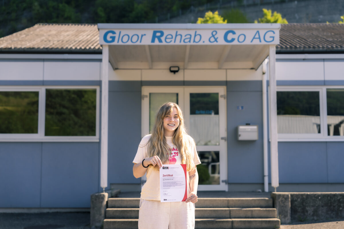 Eine lächelnde Person hält ein Zertifikat vor einem Reha-Zentrum.