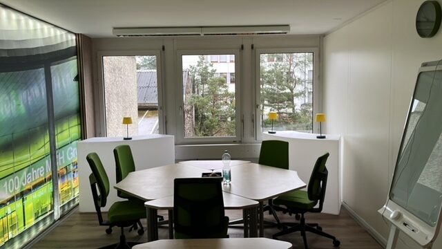 Ein heller Besprechungsraum mit Tisch, Stühlen und großen Fenstern.