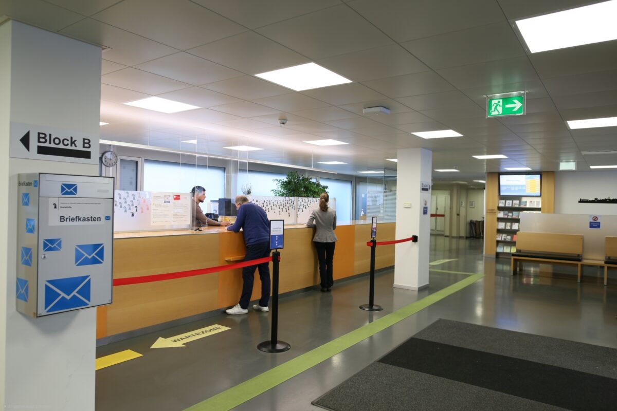 Ein Warteschlangenbereich mit zwei Personen an einem Schalter in einem Gebäude.