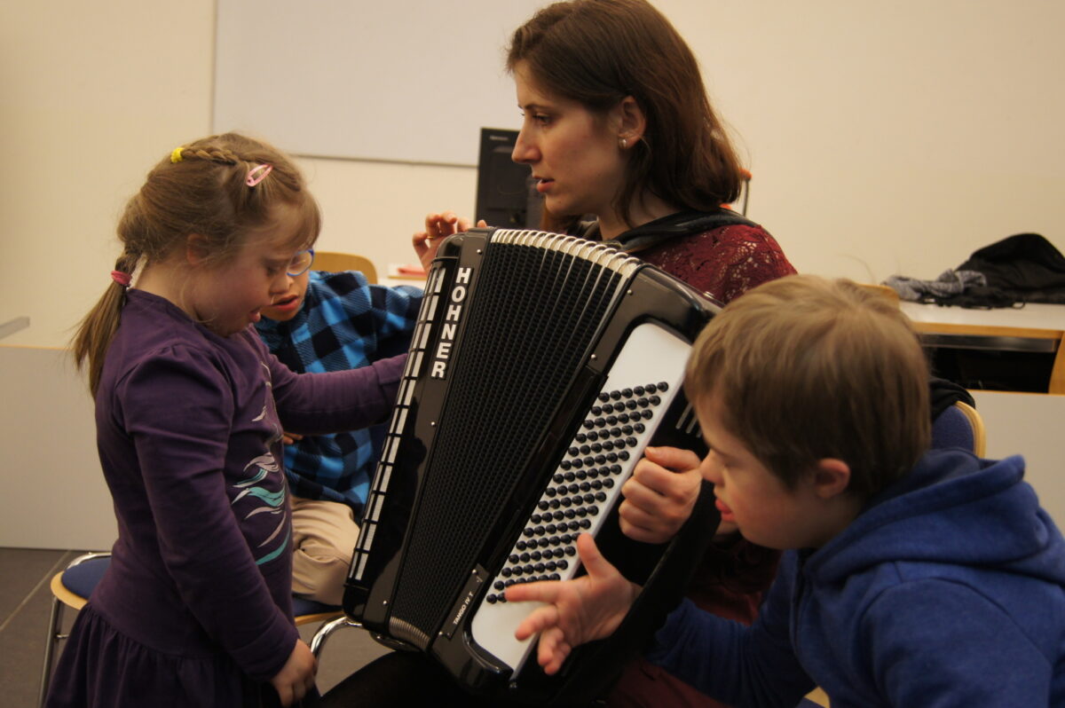 Eine Frau spielt Akkordeon mit zwei Kindern, die interessiert zuschauen.