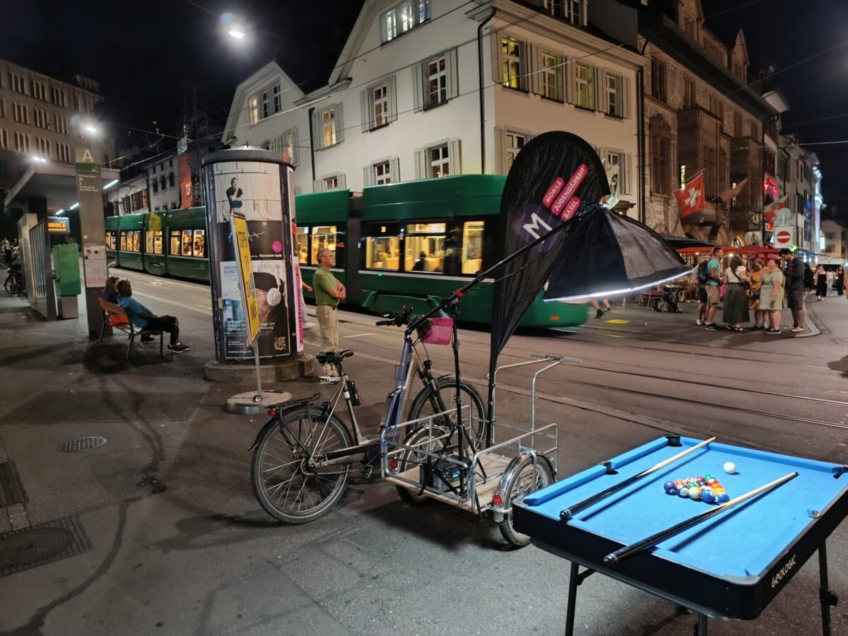 Abendliche Straßenszene mit Fahrrad, Billardtisch und vorbeifahrender Straßenbahn.