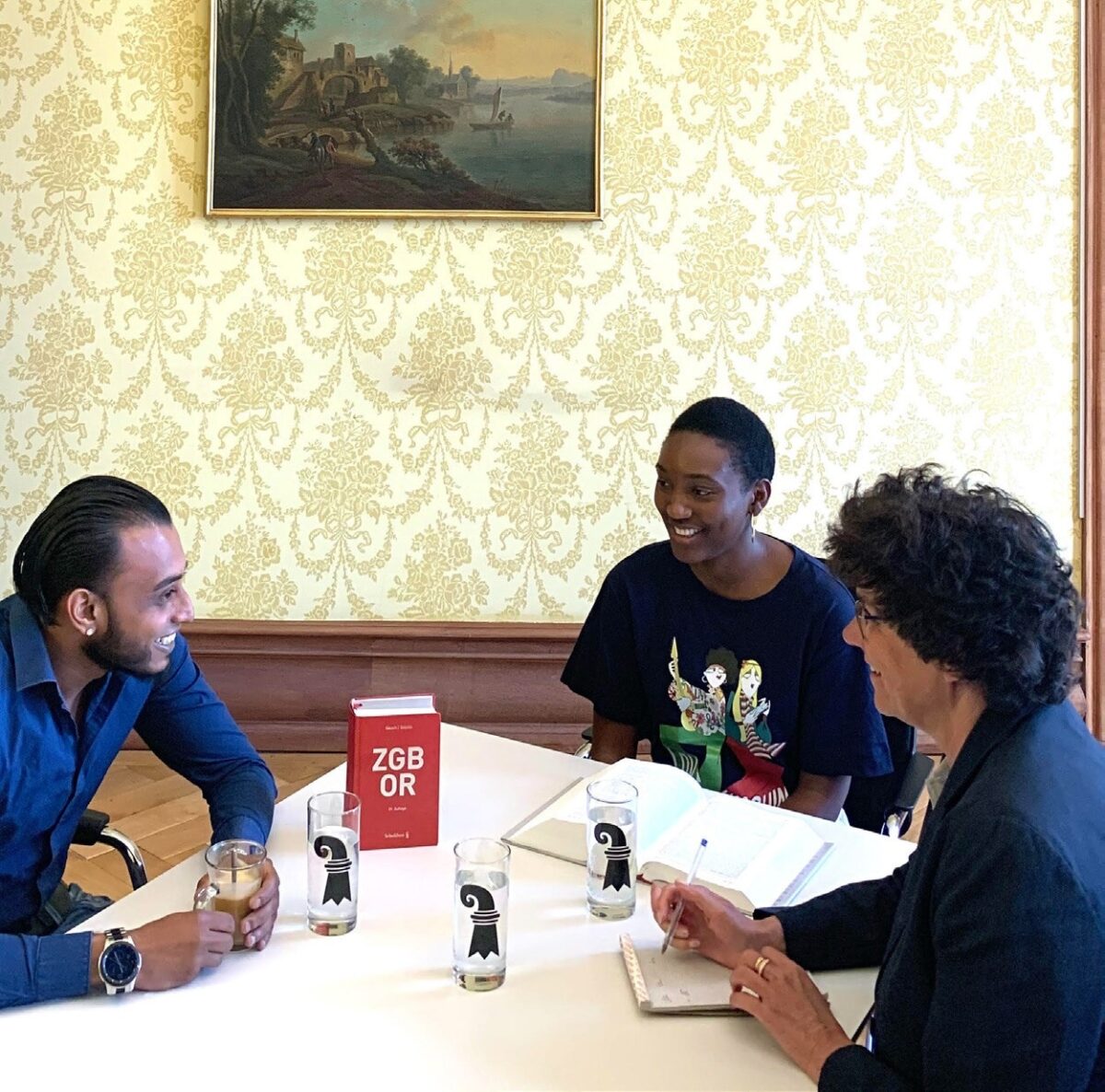 Drei Personen sitzen lachend an einem Tisch mit Gläsern und einem Buch.