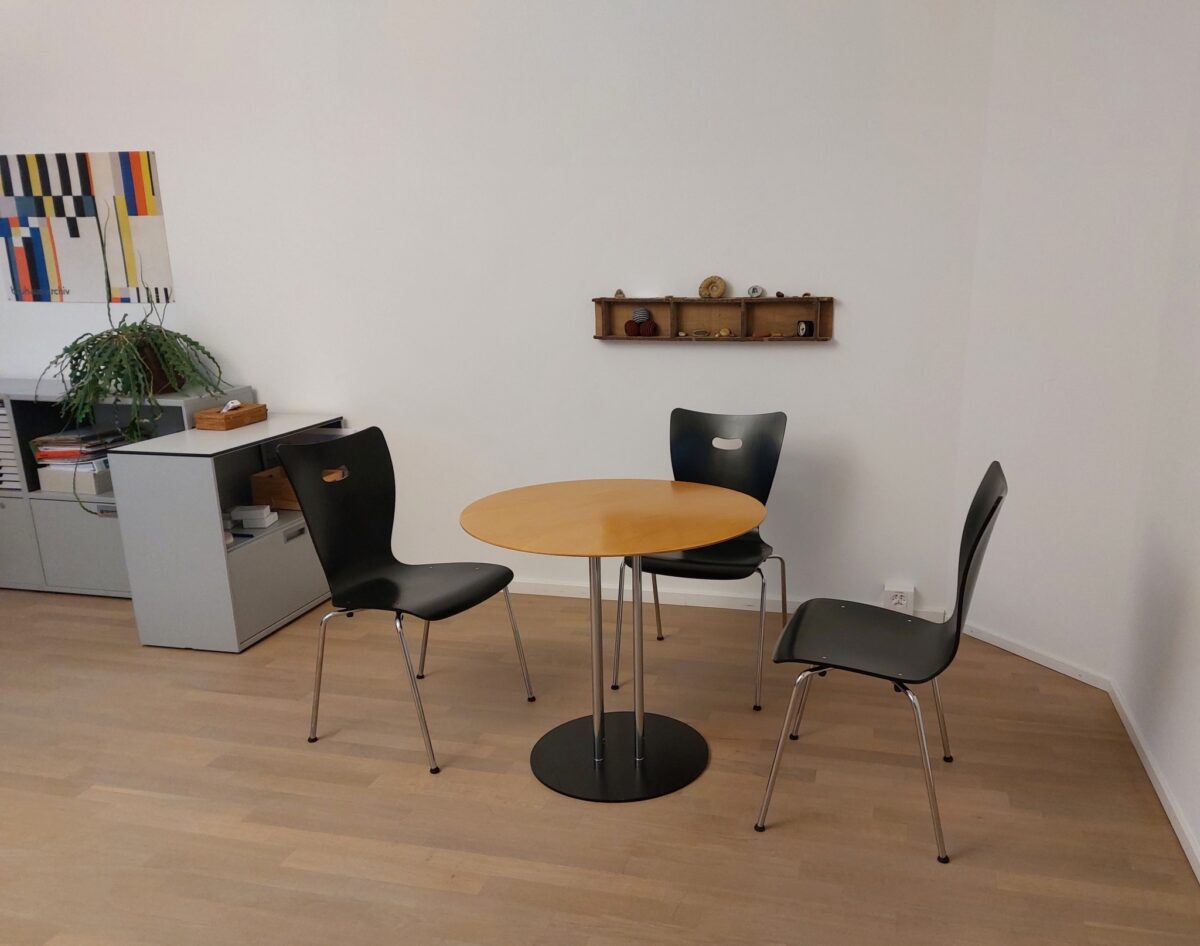 Ein kleiner Tisch mit drei Stühlen in einem hellen Raum mit Pflanzen und Regal.