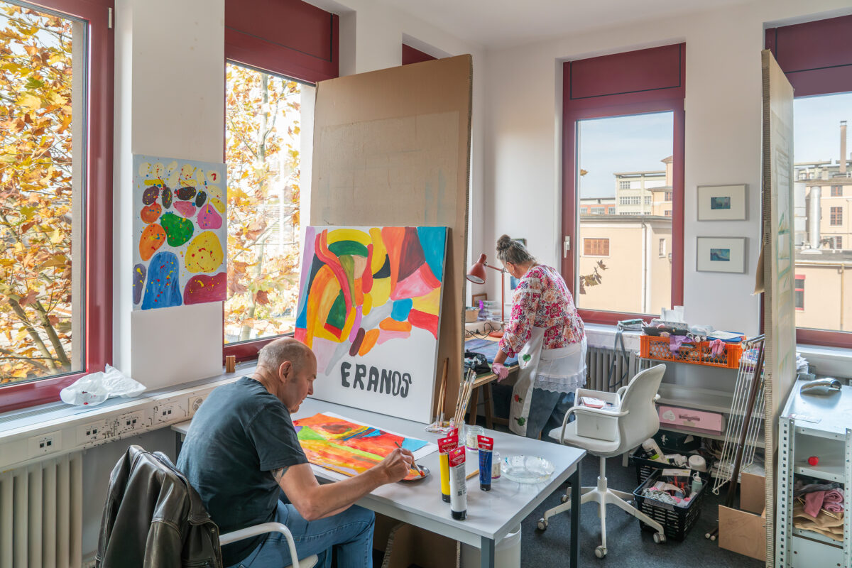 Zwei Personen malen in einem hellen Raum mit großen Fenstern bunte Bilder.