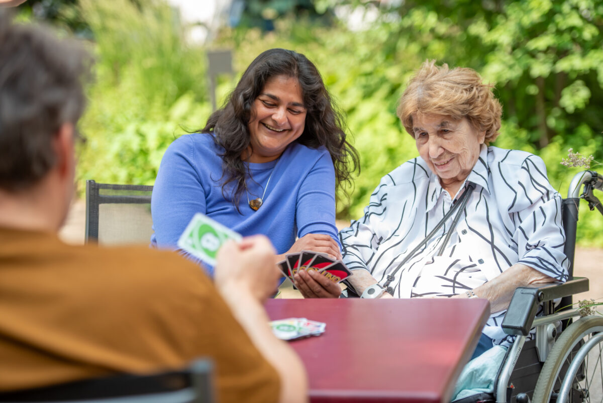 Drei Personen spielen lachend Karten im Freien, eine davon im Rollstuhl.