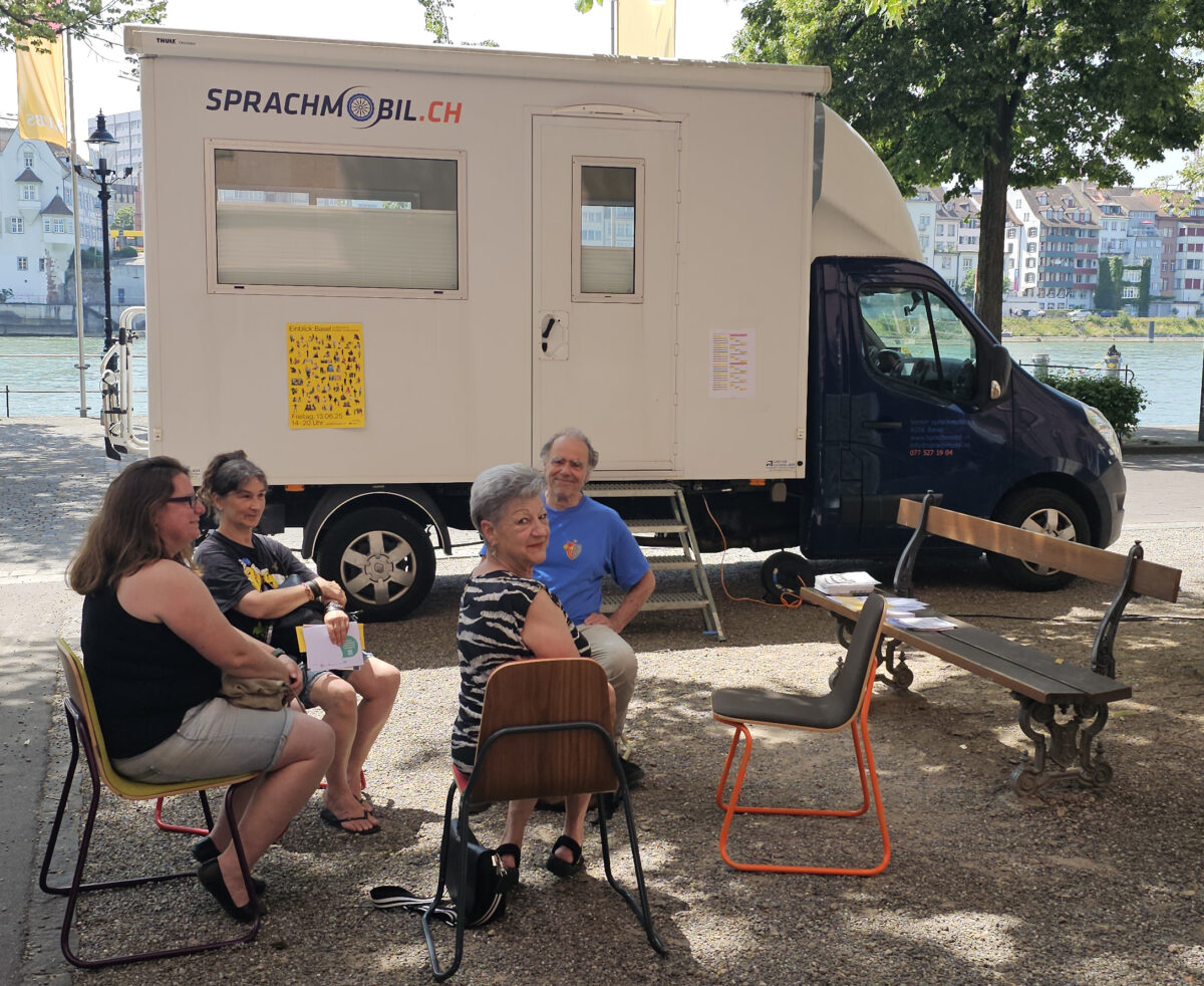 Menschen sitzen vor einem Sprachmobil am Fluss und unterhalten sich.