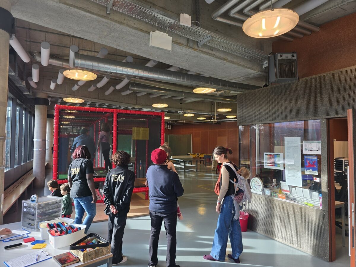 Menschen stehen in einem hellen Raum mit roten Rohren und spielen oder warten.