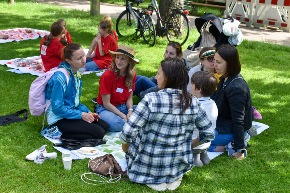 Eine Gruppe von Menschen sitzt auf einer Decke im Gras und unterhält sich.