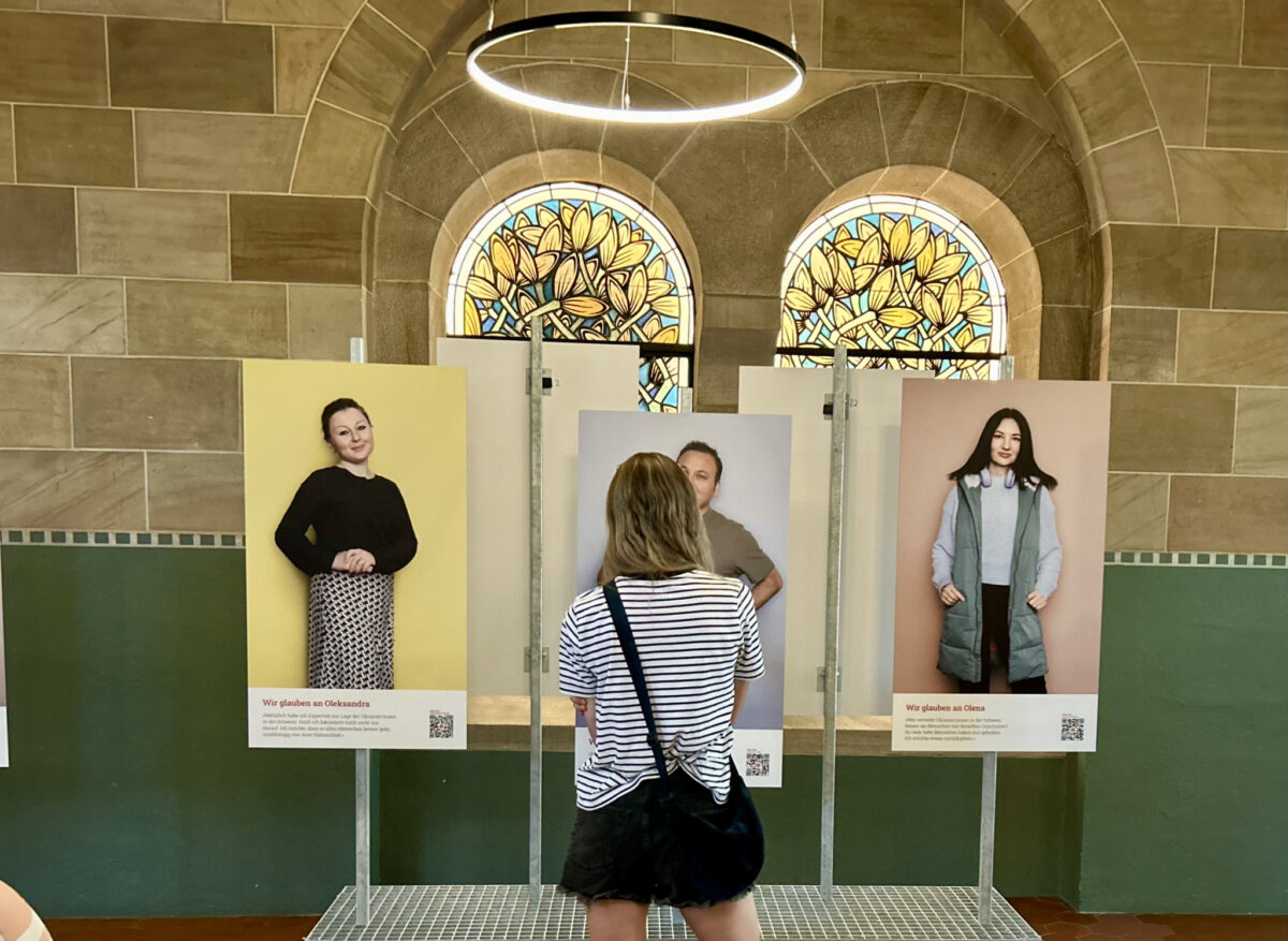 Zwei Personen betrachten Ausstellungsplakate in einem Raum mit farbigen Fenstern.