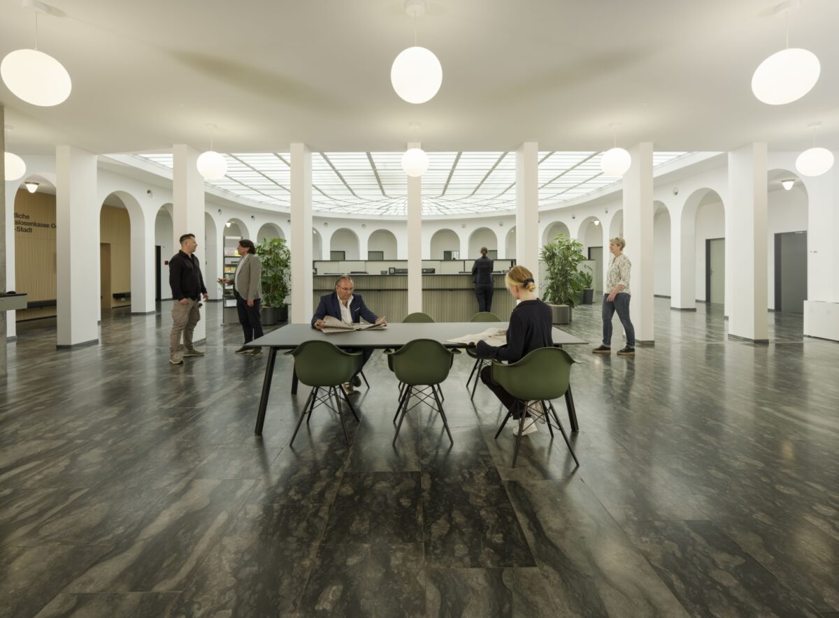 Menschen sitzen und stehen in einem hellen, modernen Innenhof mit Arkaden und Pflanzen.