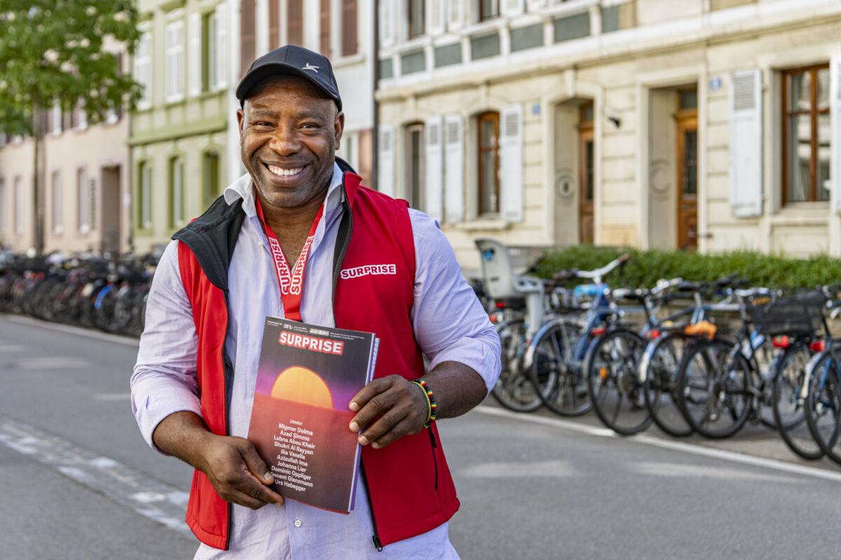 Ein lächelnder Mann hält ein Magazin mit dem Titel "Surprise" auf einer Straße.