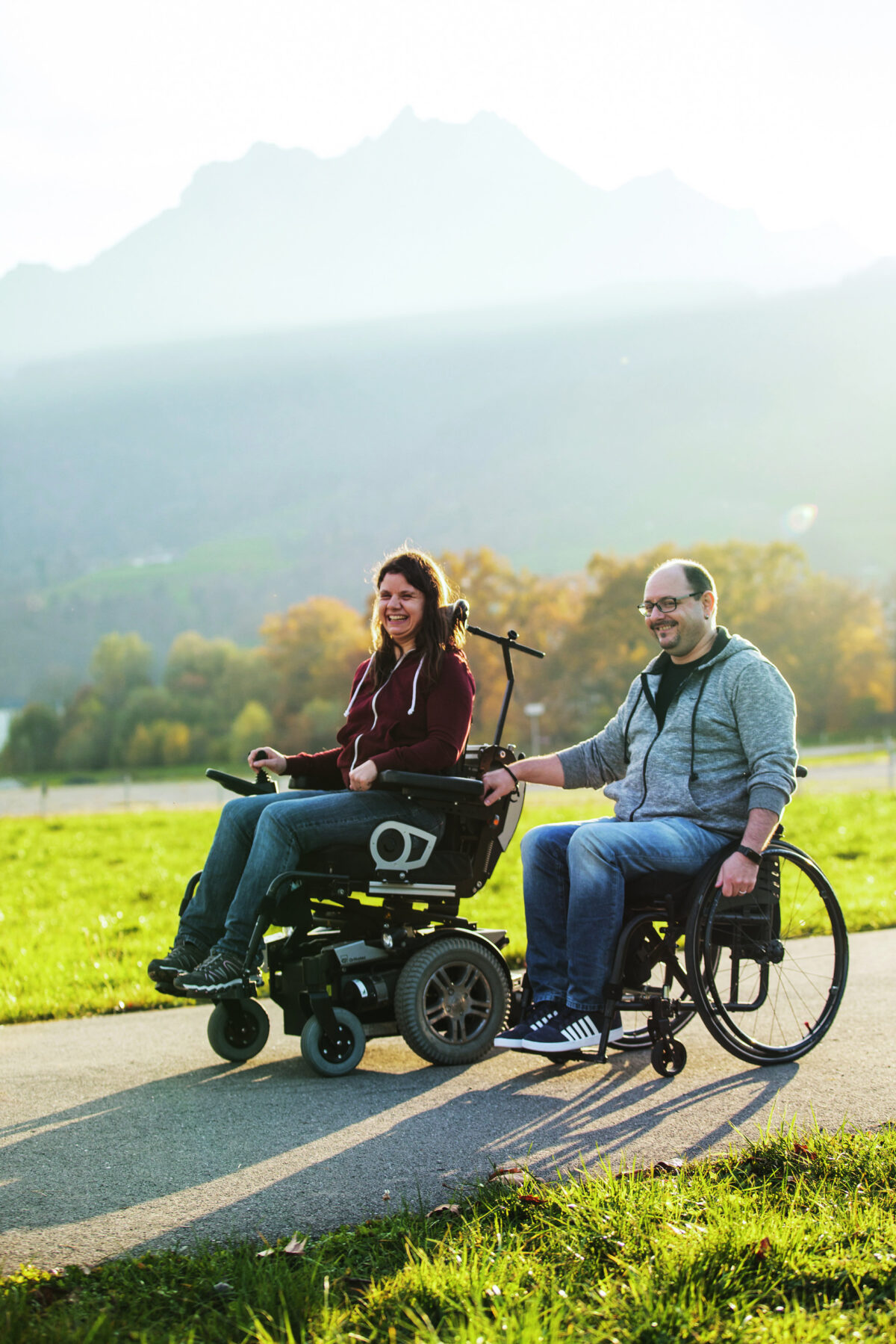 Zwei Personen lachen in ihren Rollstühlen auf einem Weg in der Natur.