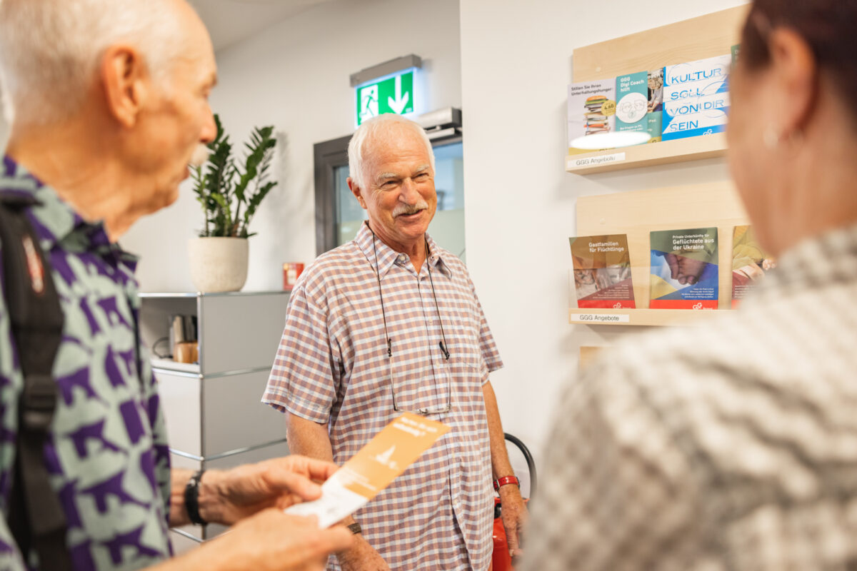 Drei ältere Menschen unterhalten sich in einem Raum mit Informationspostern.