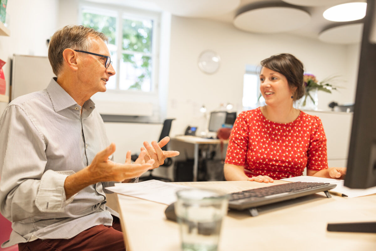 Zwei Personen unterhalten sich in einem Büro an einem Tisch mit Dokumenten.