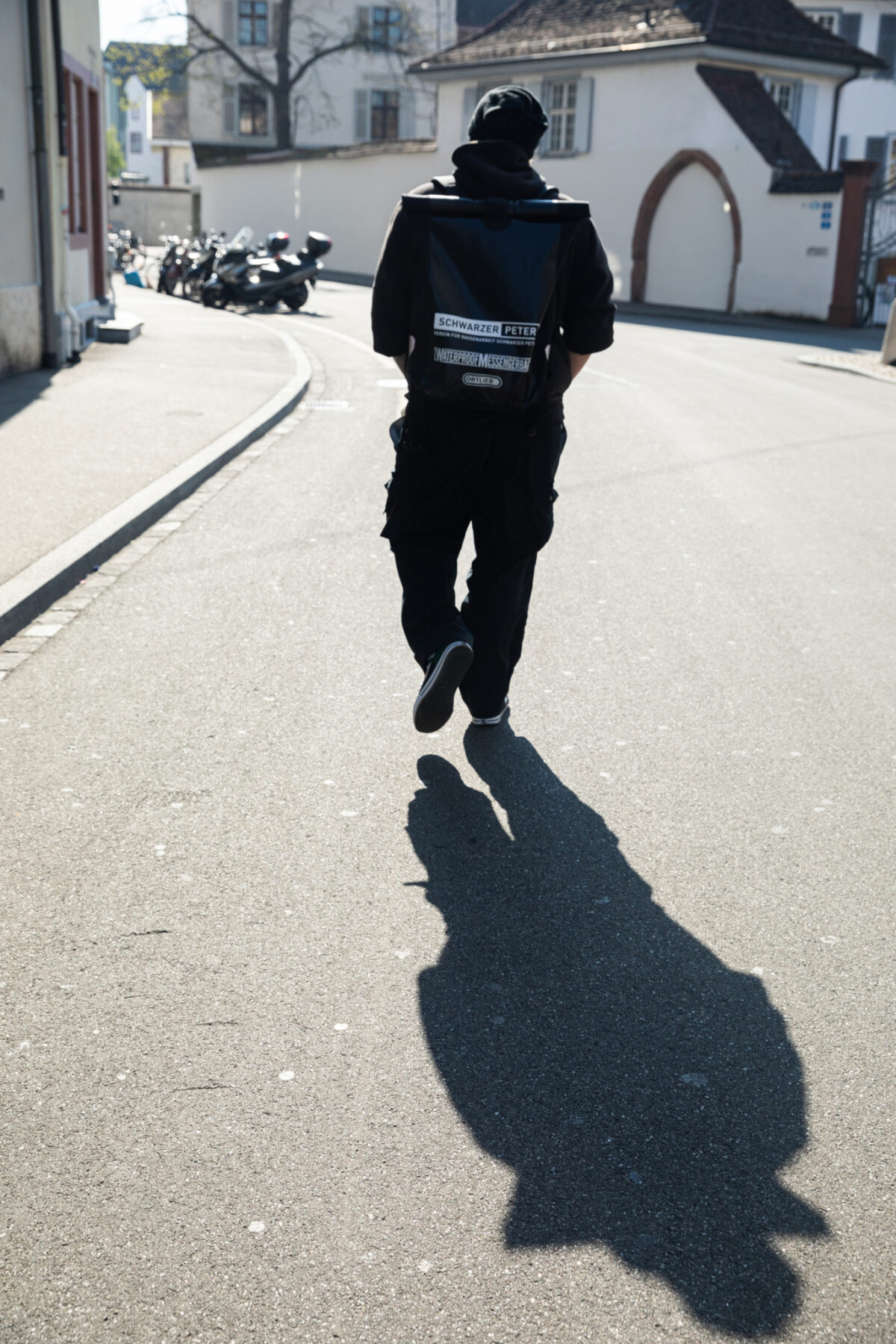 Eine Person in schwarzer Kleidung geht eine Straße entlang und trägt eine große Tasche.