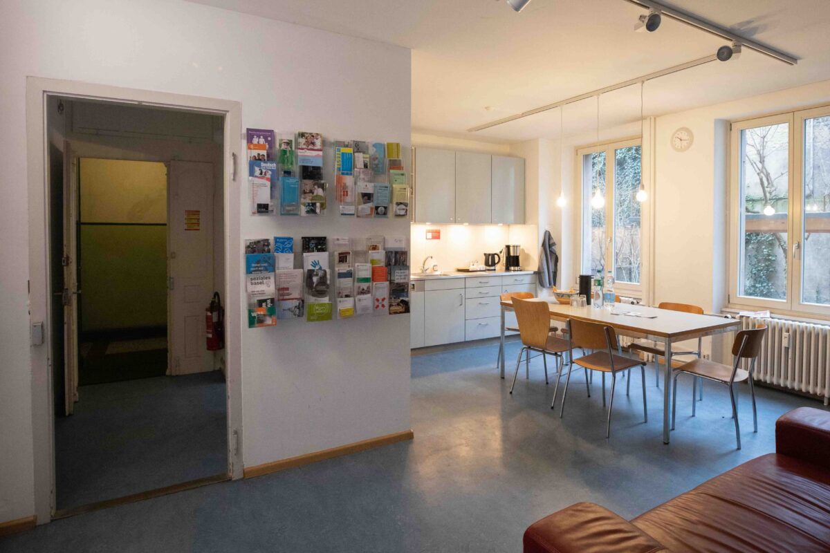 Ein heller Raum mit Küche, Tisch, Stühlen und Informationsmaterial an der Wand.