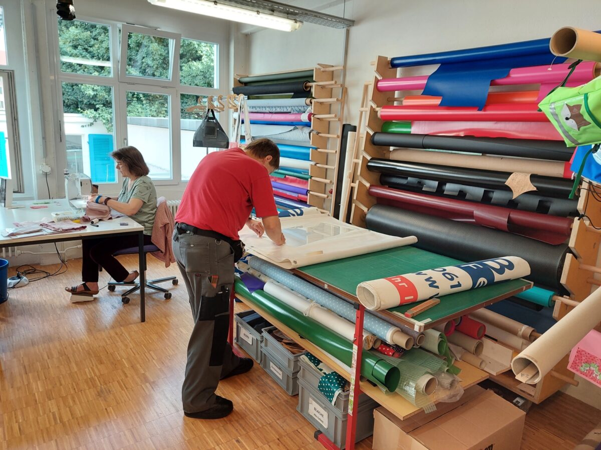 Zwei Personen arbeiten in einem Raum mit Stoffrollen und Materialien.