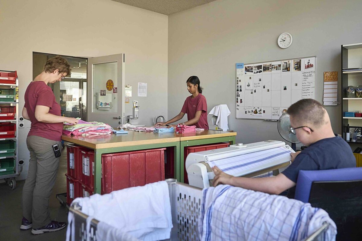 Drei Personen falten in einem Raum Wäsche und sortieren sie in Schubladen.