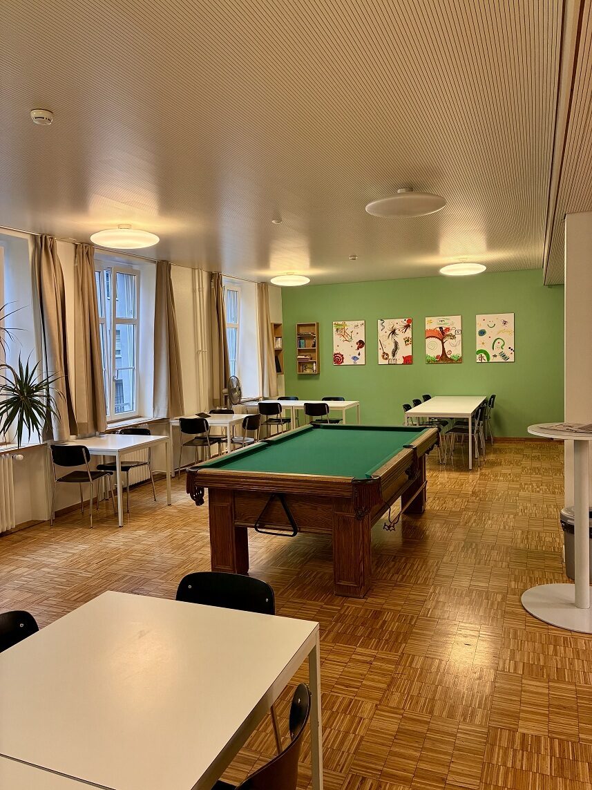 Ein Aufenthaltsraum mit einem Billardtisch. Im Raum stehen auch Tische mit Stühlen darum. Es gibt eine grüne Wand. An der Wand hängen vier Bilder und ein kleines Bücherregal.