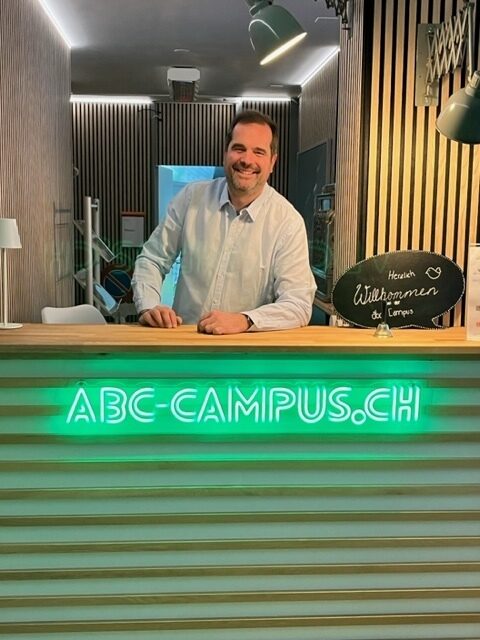 Ein lächelnder Mann steht hinter einem Empfangstresen mit der Aufschrift "ABC-CAMPUS.CH".