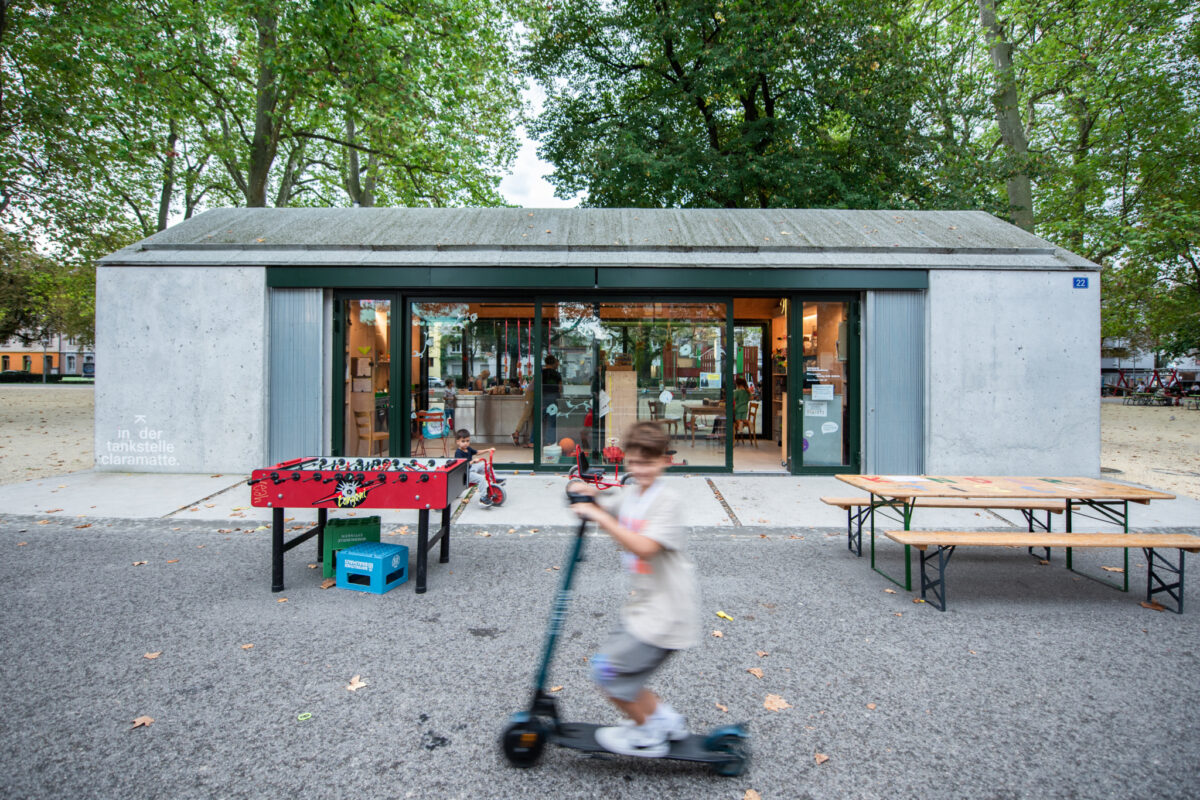 Ein Kind fährt mit einem Tretroller vor einem kleinen Gebäude mit offenen Türen.