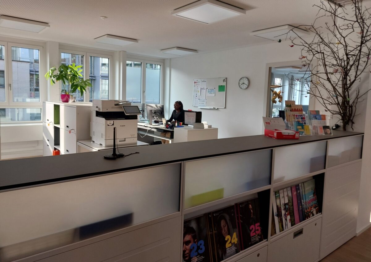 Es ist ein Empfangsbereich zu sehen. Im Hintergrund arbeitet eine Frau und Pflanzen sind zu sehen.