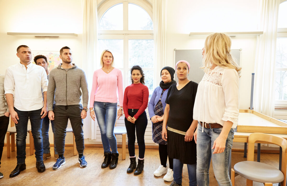 Eine Gruppe von Menschen steht in einem hellen Raum und hört einer Frau zu.