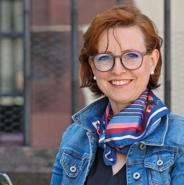 Eine lächelnde Frau mit Brille und buntem Schal vor einem Gebäude.