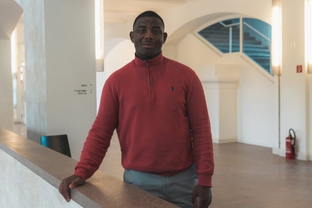 Ein Mann in rotem Pullover lehnt an einem Geländer in einem hellen Gebäude.