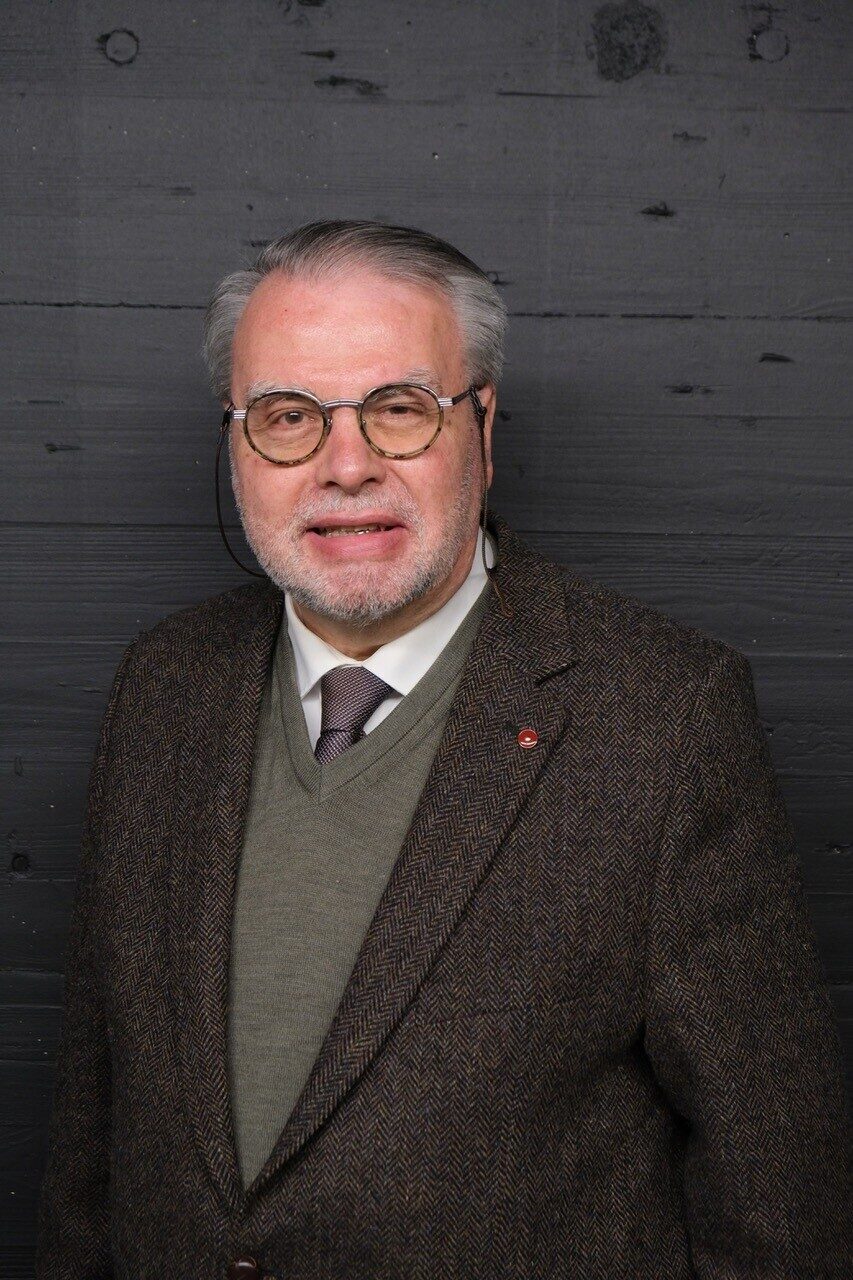 Ein älterer Mann mit Brille und Anzug steht vor einer dunklen Holzwand.