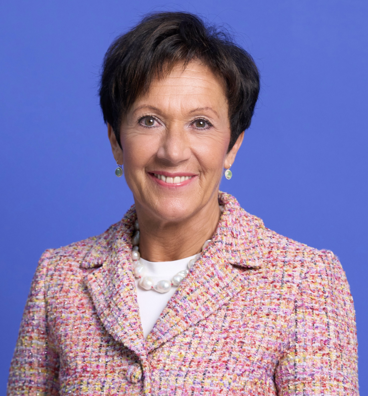 Eine lächelnde Frau mit kurzem Haar in einem bunten Blazer vor blauem Hintergrund.