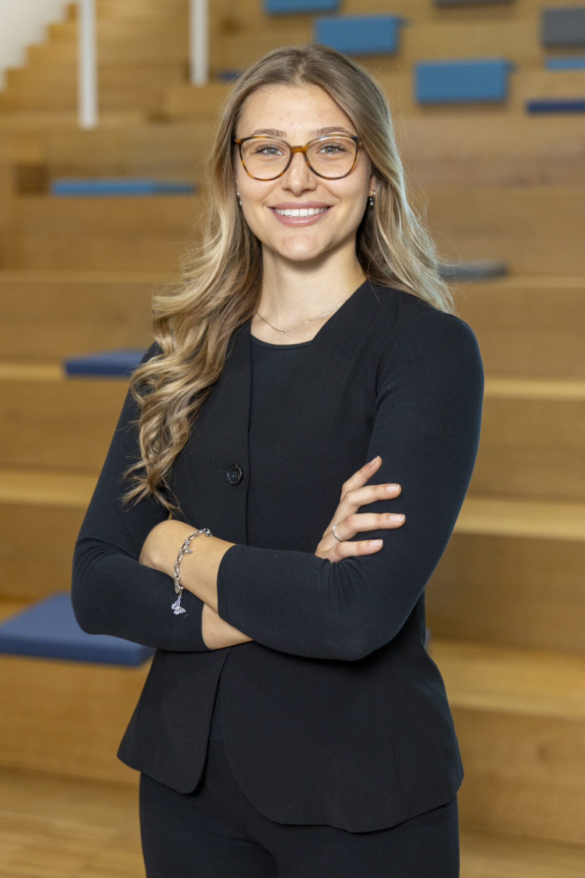 Eine lächelnde Frau mit Brille steht mit verschränkten Armen in einem Raum mit Holzvertäfelung.