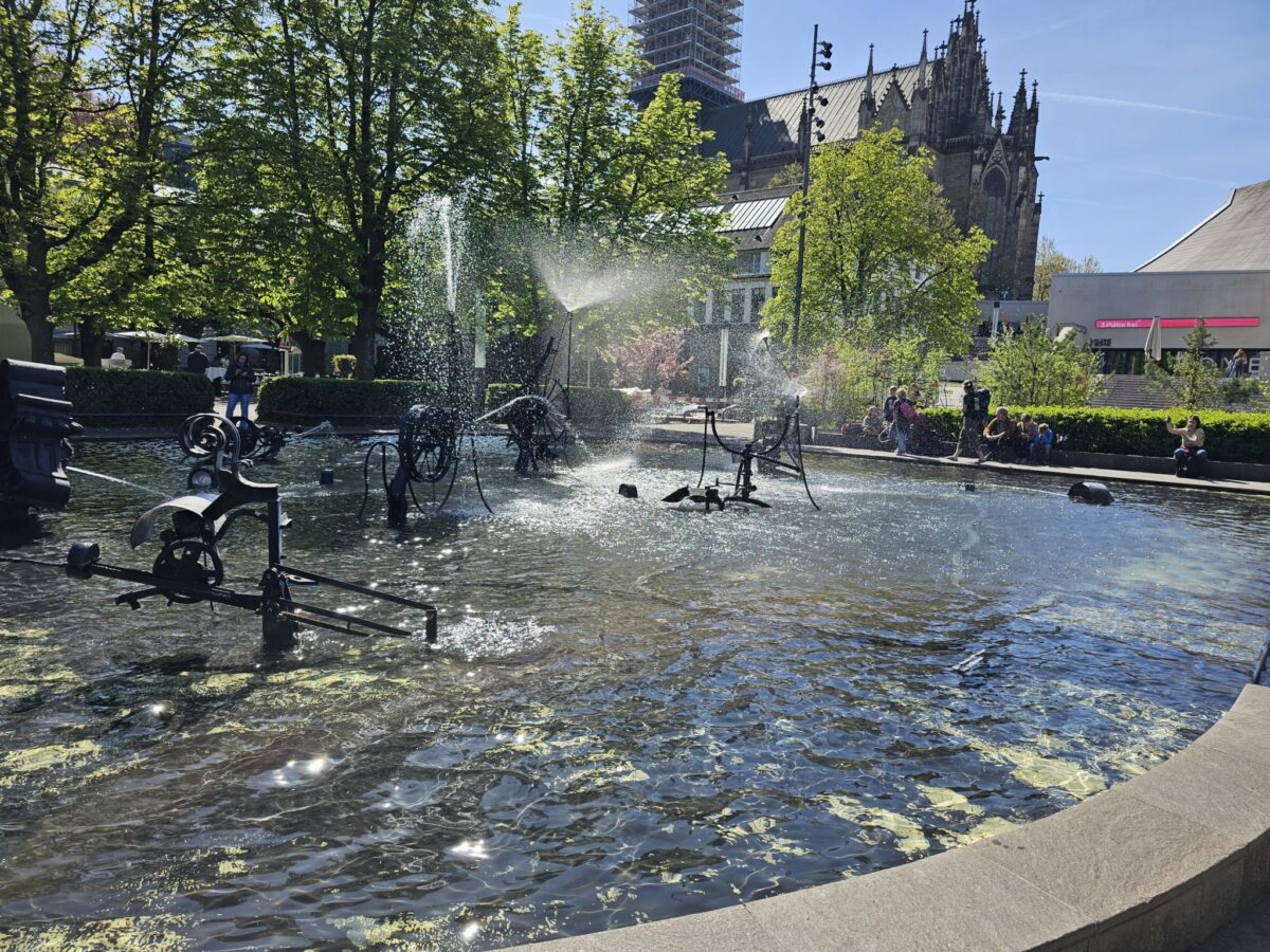 Ein Brunnen mit Wasserspiel und alten Fahrrädern im Vordergrund, im Hintergrund eine gotische Kirche.