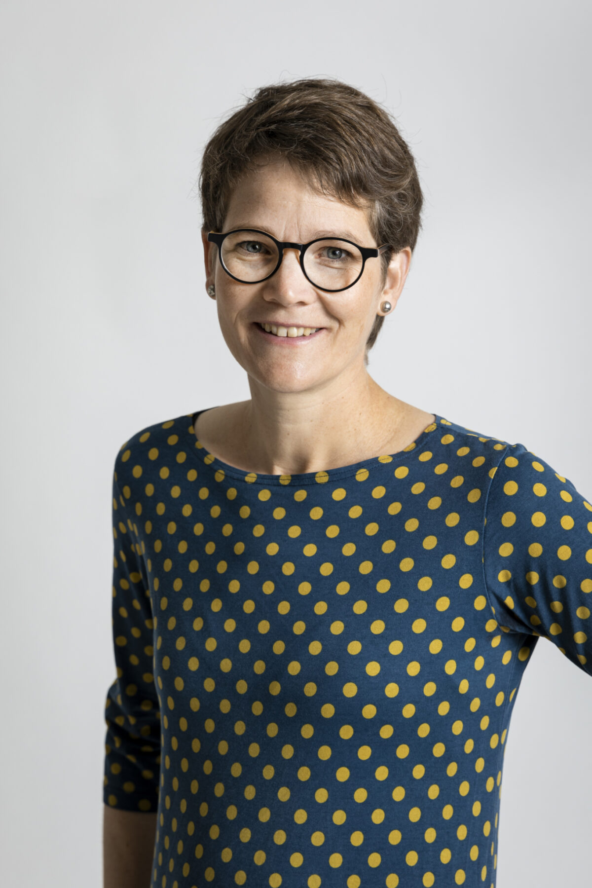 Eine lächelnde Frau mit kurzen Haaren und Polka-Dot-Shirt trägt eine Brille.