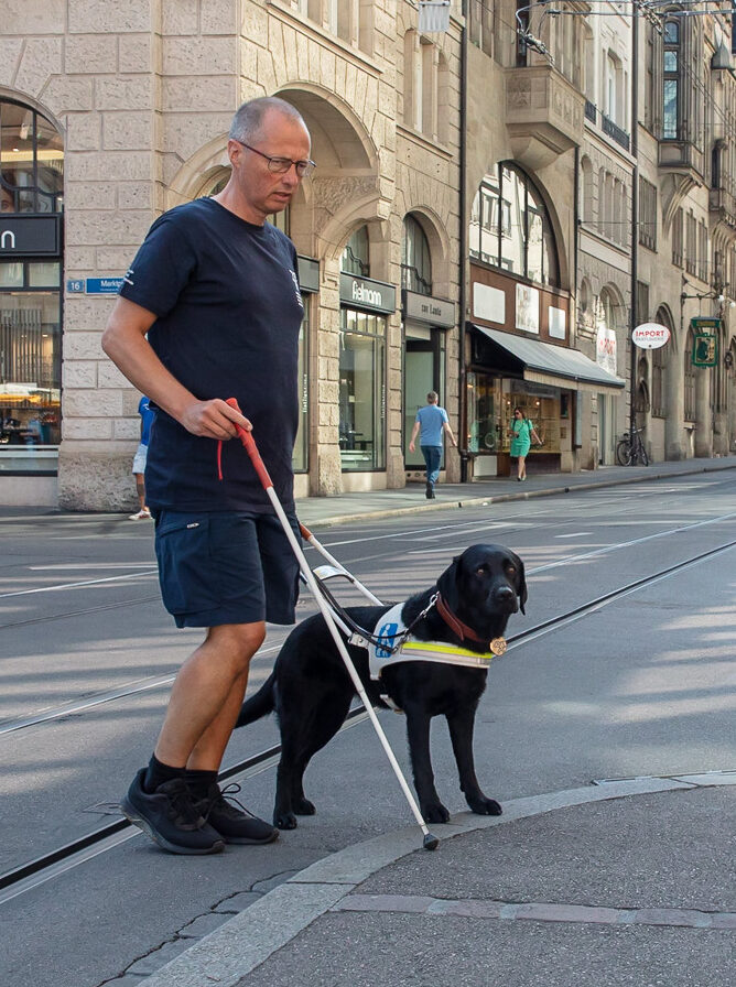Ein Mann mit einem Blindenhund überquert eine Straße in einer Stadt.