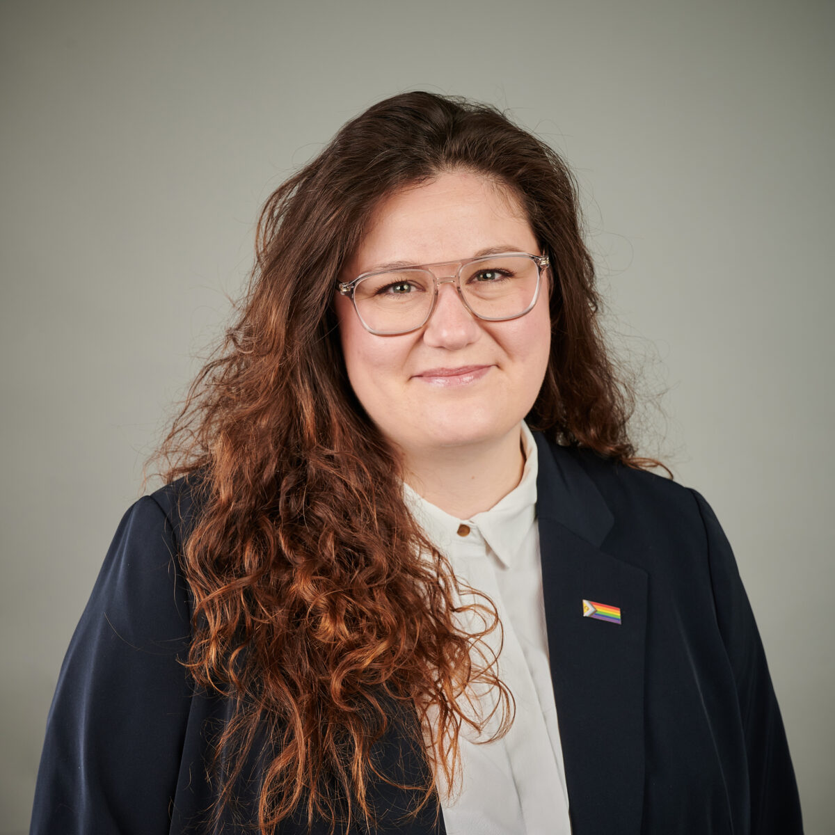 Eine Person mit Brille und Regenbogen-Anhänger lächelt in die Kamera.